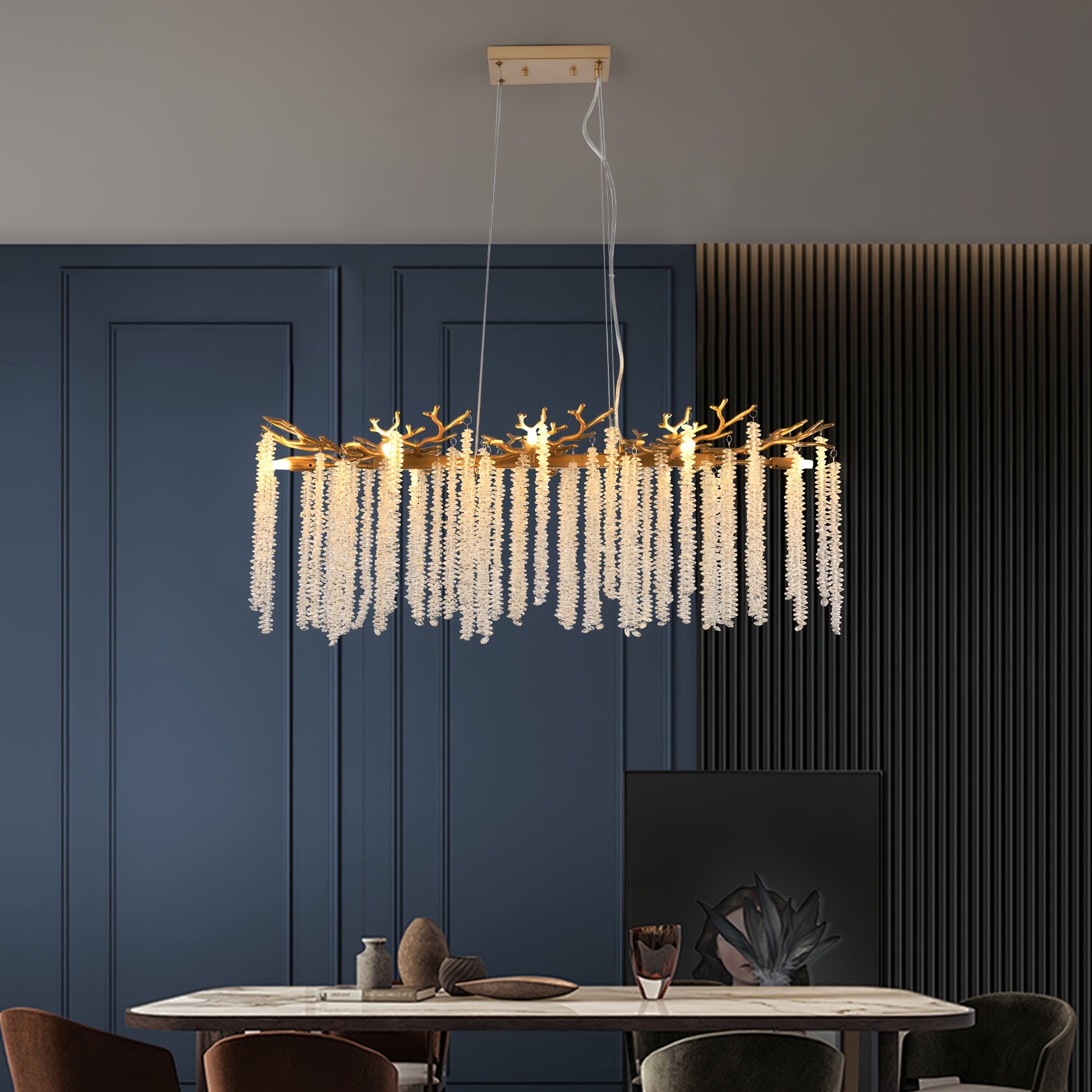 Suspended light with gold branch frame and crystal drops, positioned above a marble dining table with blue wall and dark chairs