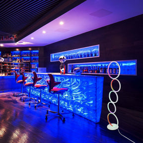 Vibrant bar interior featuring a circular floor lamp with glowing rings, placed beside a color-lit counter and red bar stools