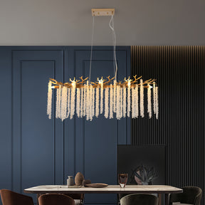 Suspended light with gold branch frame and crystal drops, positioned above a marble dining table with blue wall and dark chairs