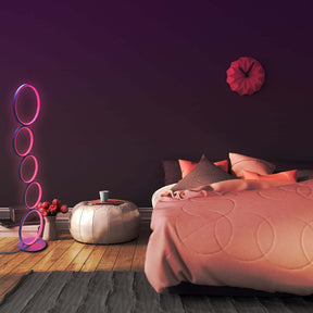 Cozy bedroom scene featuring an RGB circular floor lamp with pink glowing rings, placed beside a low stool and flower vase on a wooden floor